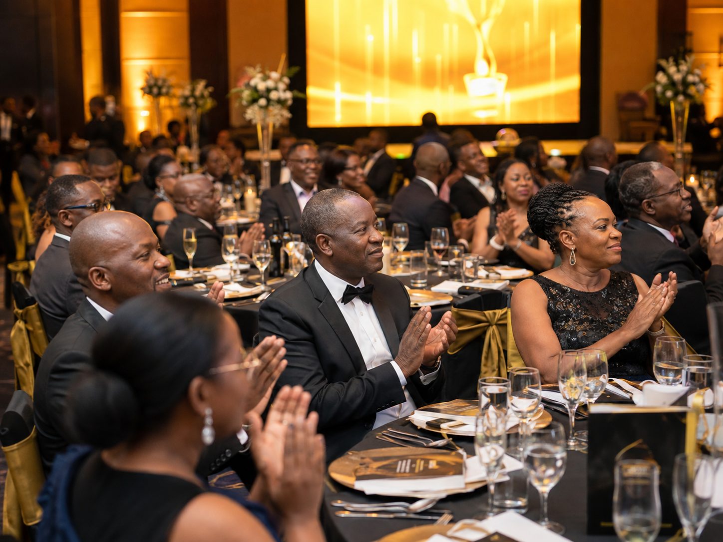 Guests in formal evening wear applauding at round banquet tables during an awards dinner, stage lit in warm gold behind them