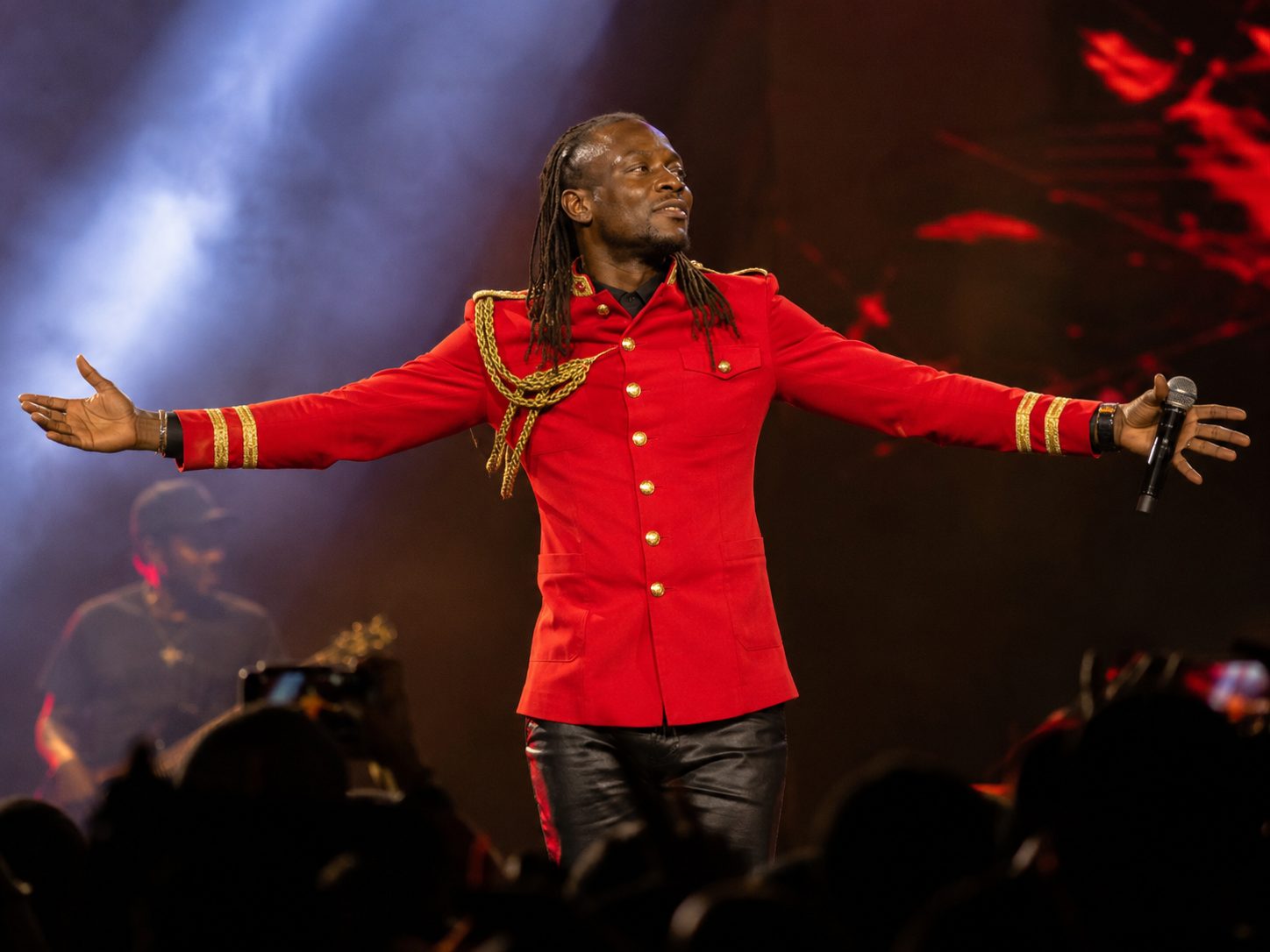 Jah Prayzah performing on stage in a red military-style jacket, arms outstretched, crowd silhouetted in front of the stage
