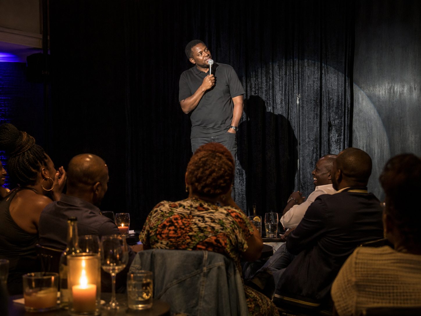 A stand-up comedian mid-performance under a single spotlight, audience laughing at candlelit tables in the foreground