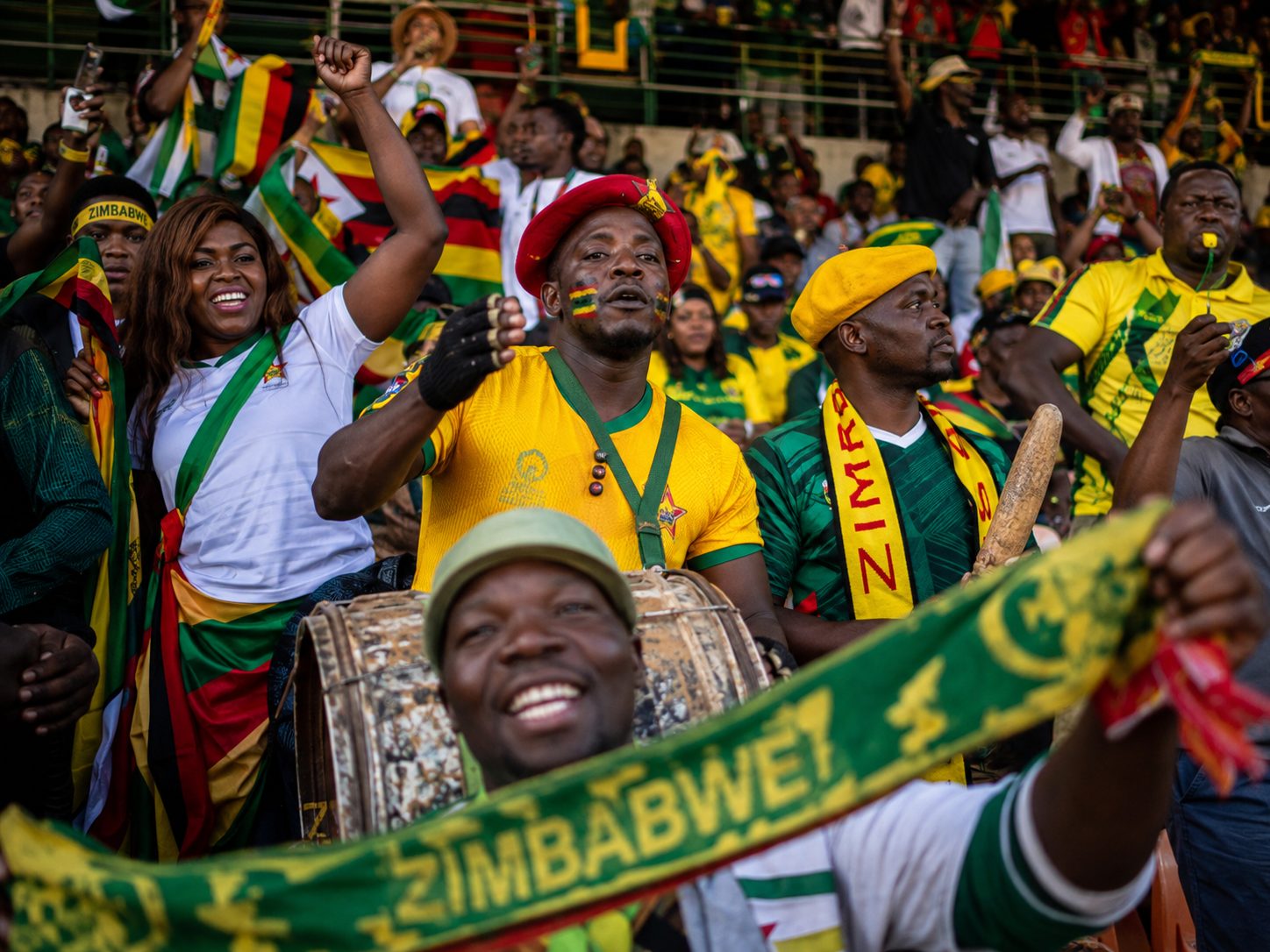 Football supporters in yellow and green Zimbabwe colours celebrating in a packed stadium stand, drums and flags visible