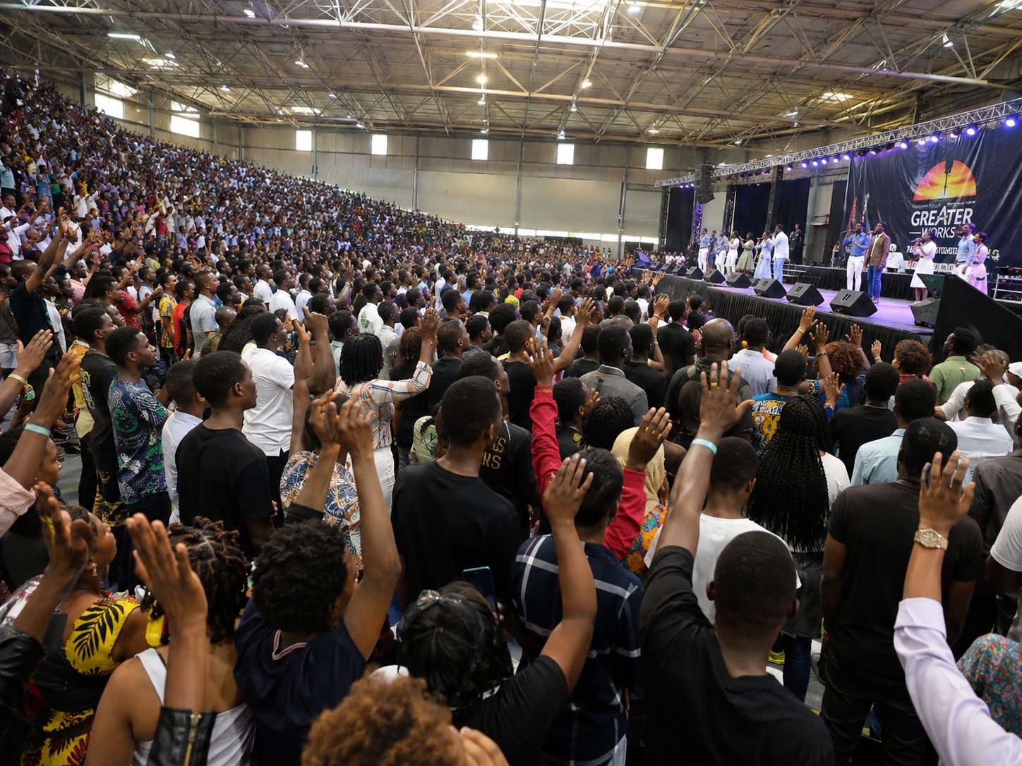 A large church conference audience standing with hands raised in a filled indoor arena, worship team visible on stage