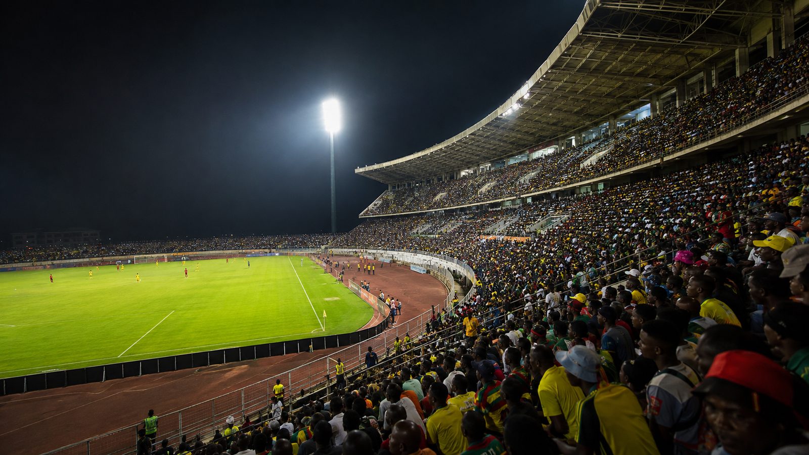Floodlit national stadium at night packed with supporters, green pitch bright against the dark stands